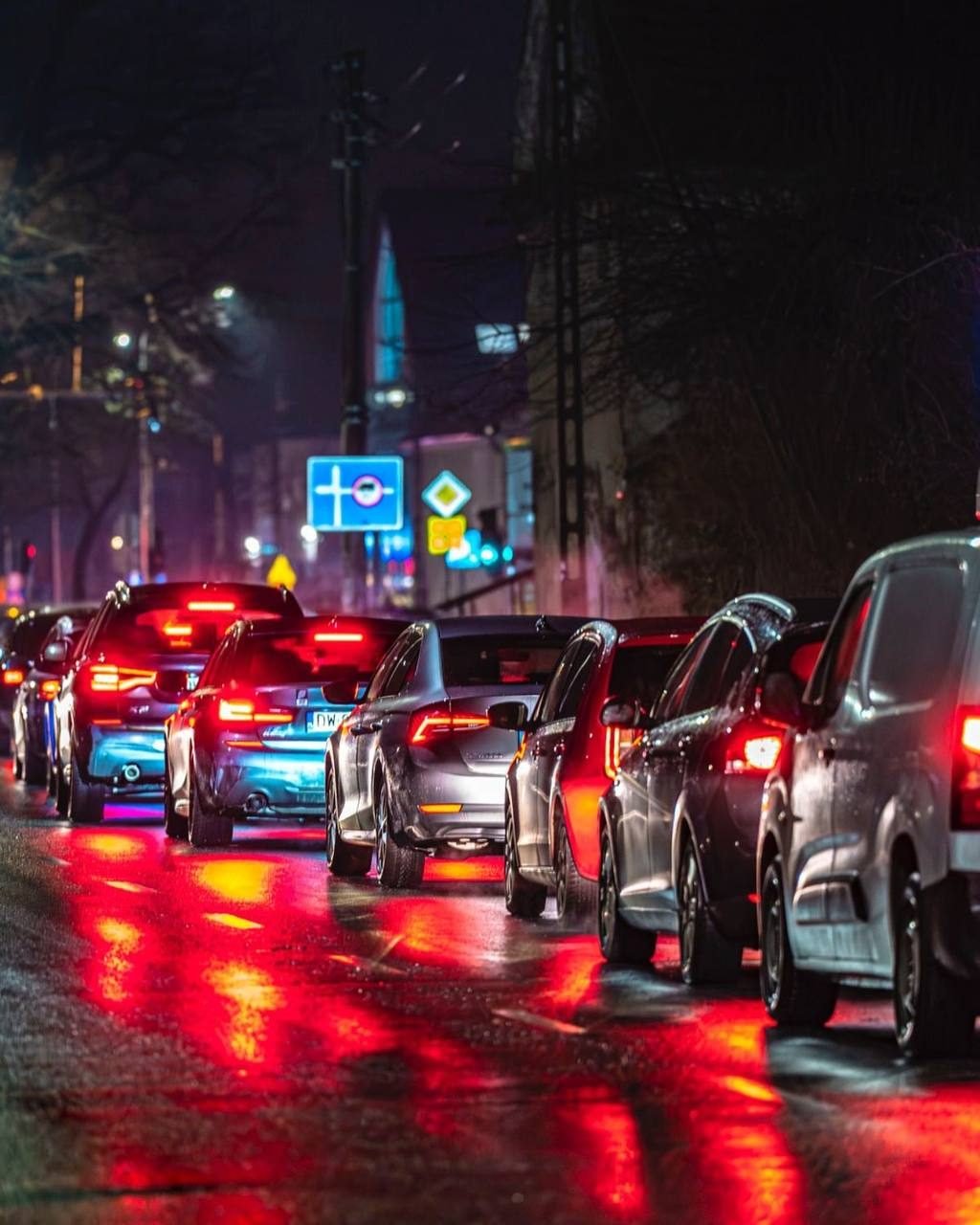 Fila de carros con luces traseras rojas reflejadas en el pavimento mojado de noche